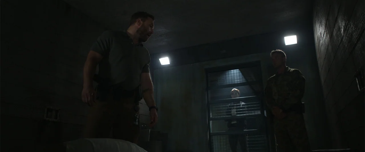 Black Site (2022) movie still: medium — A dimly lit industrial room contains concrete walls and a metal cell gate. Two men in the foreground stand…