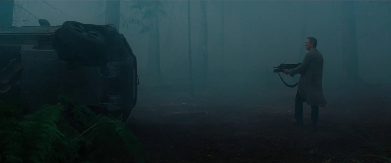 No Time To Die (2021) movie still: wide — A dense forest clearing filled with heavy fog and tall trees. A man holds a rifle while standing next to a…