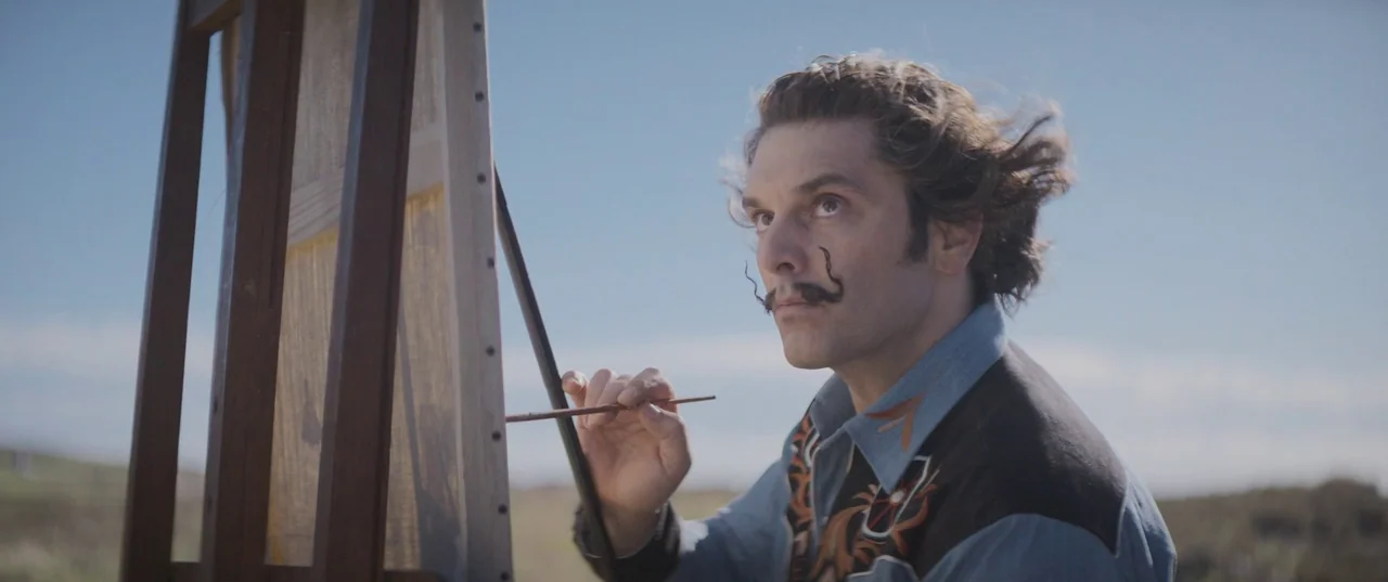 Daaaaaalí! (2023) movie still: medium — A man stands outdoors in a wide open area before a wooden easel. He holds a thin brush against the canvas…