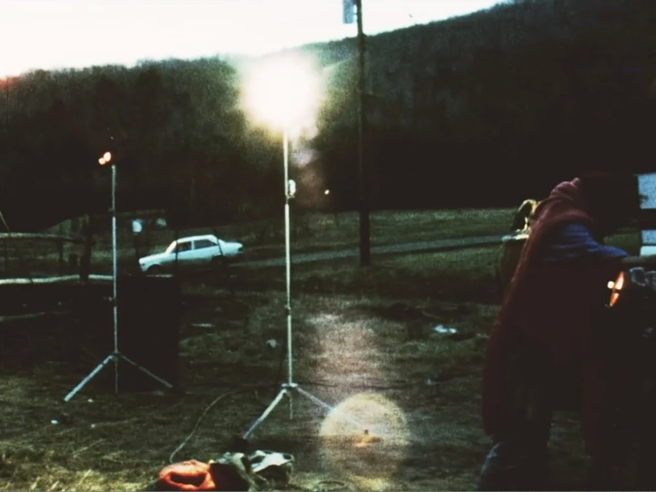 Don't Expect Too Much (2011) movie still: wide — An open grassy field at night illuminated by a single portable light stand. A person viewed from…