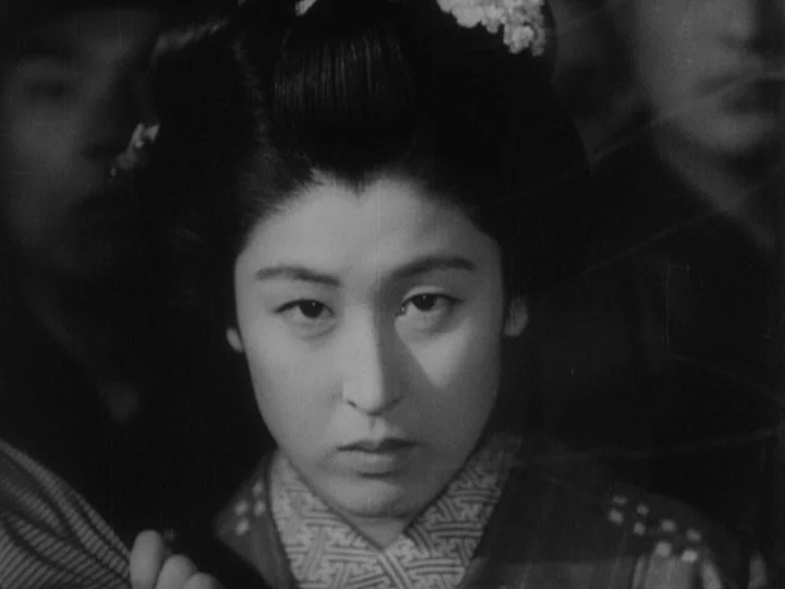 Sanshiro Sugata (1943) movie still: closeup — A woman stands in an indoor space with blurred figures visible in the background. The focus remains…