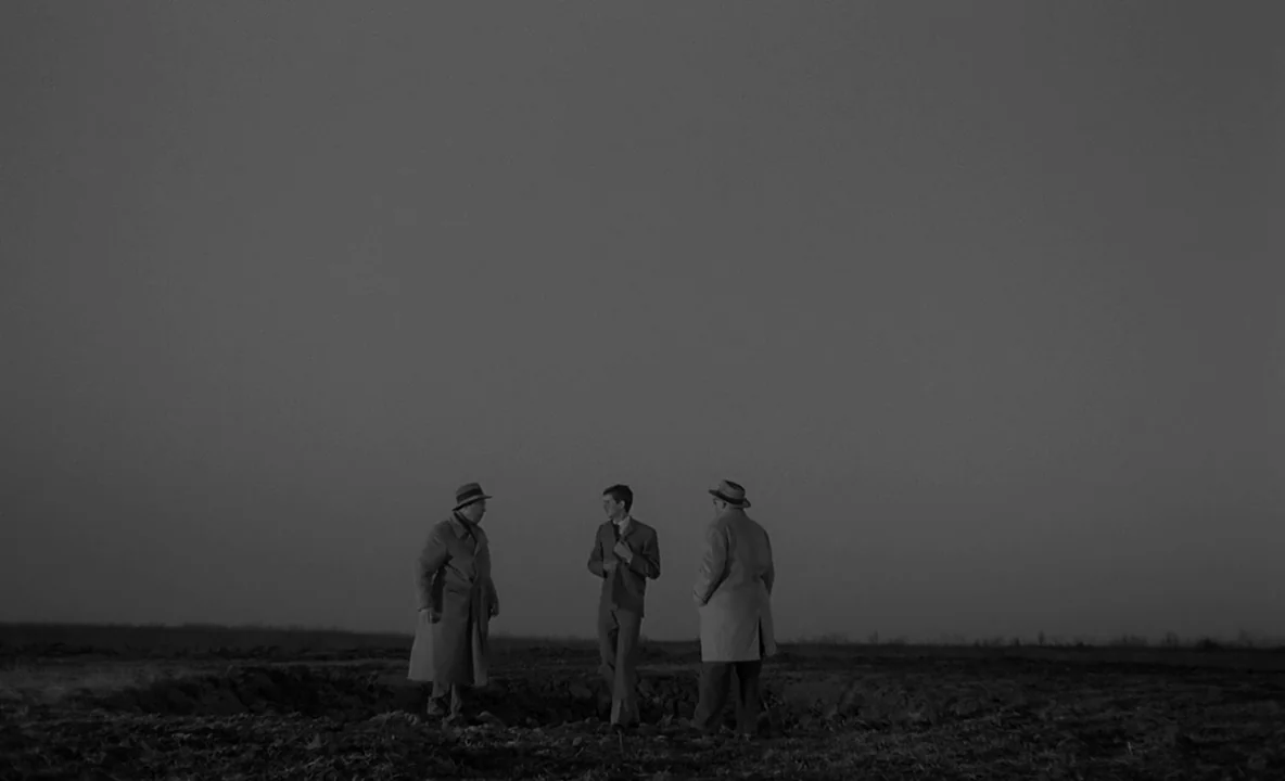 The Trial (1962) movie still: wide — Three men stand in a vast, desolate field under a dim, overcast sky. The men talk to each other while wearing…