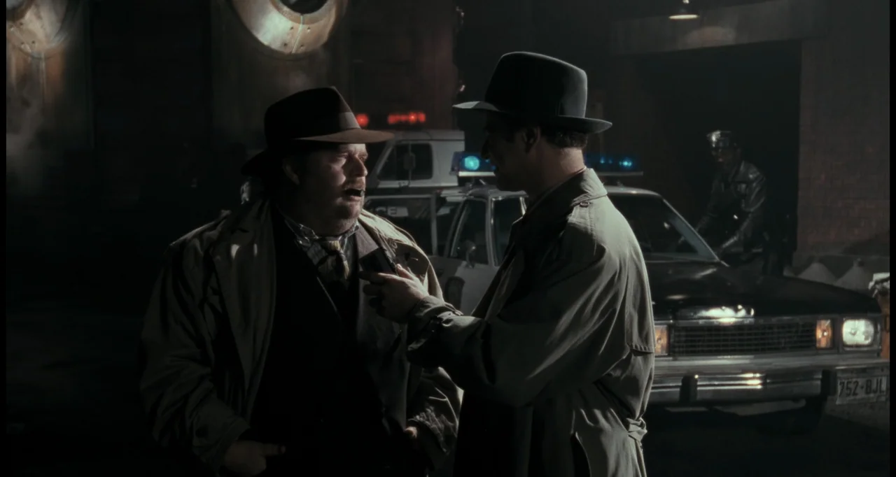 Batman (1989) movie still: medium — A dark night scene at a paved lot with a parked police cruiser behind subjects. Two men in trench coats and…