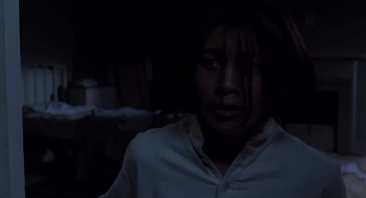 Martyrs (2008) movie still: medium — A dark bedroom with an obscured bed visible in the blurred background. A young person in a light shirt stands…