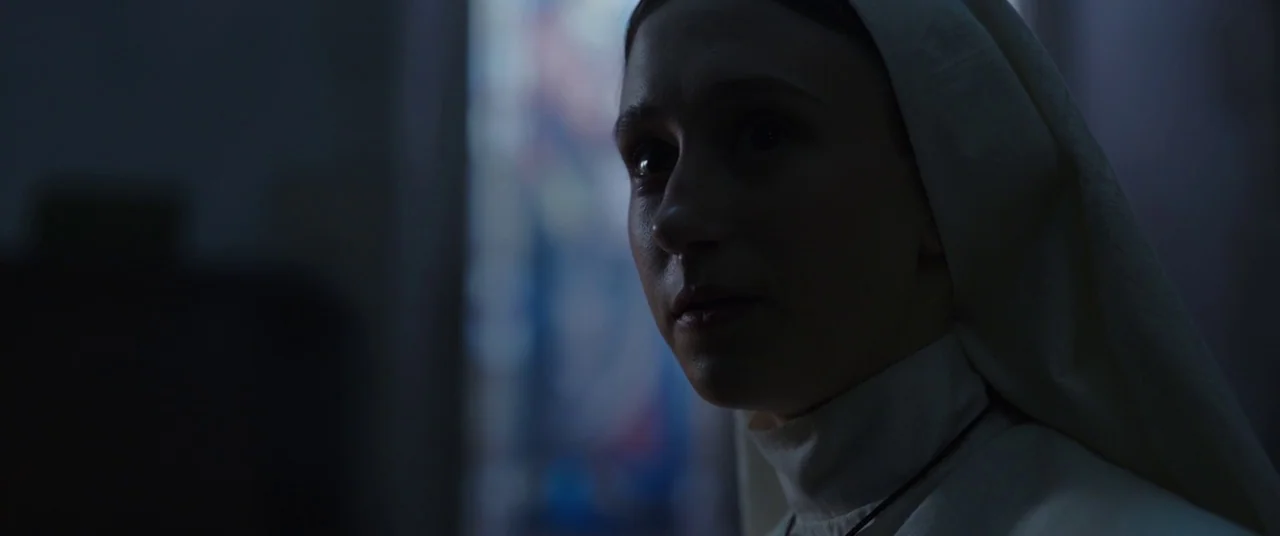 The Nun (2018) movie still: closeup — A dimly lit room features a nun positioned near a window. The subject turns her head slightly while looking…