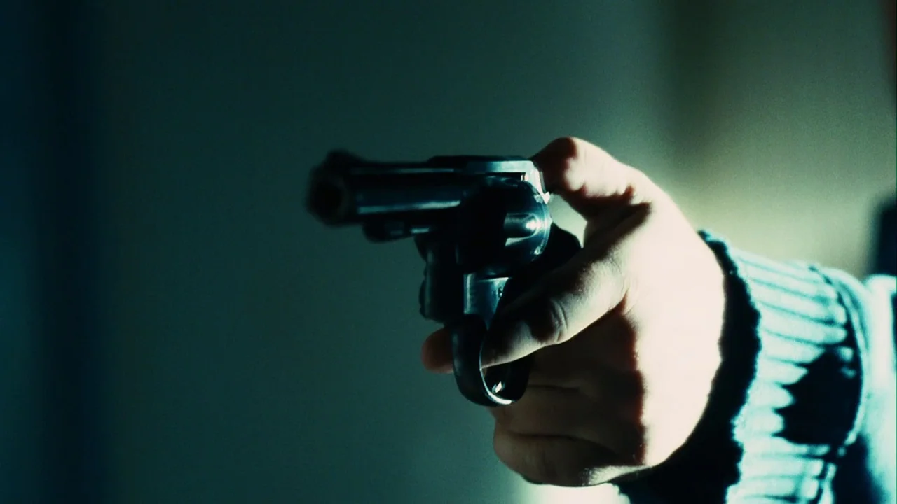 Saw III (2006) movie still: closeup — A close-up captures a dark revolver gripped by a person's hand. The background is a solid, out-of-focus muted…