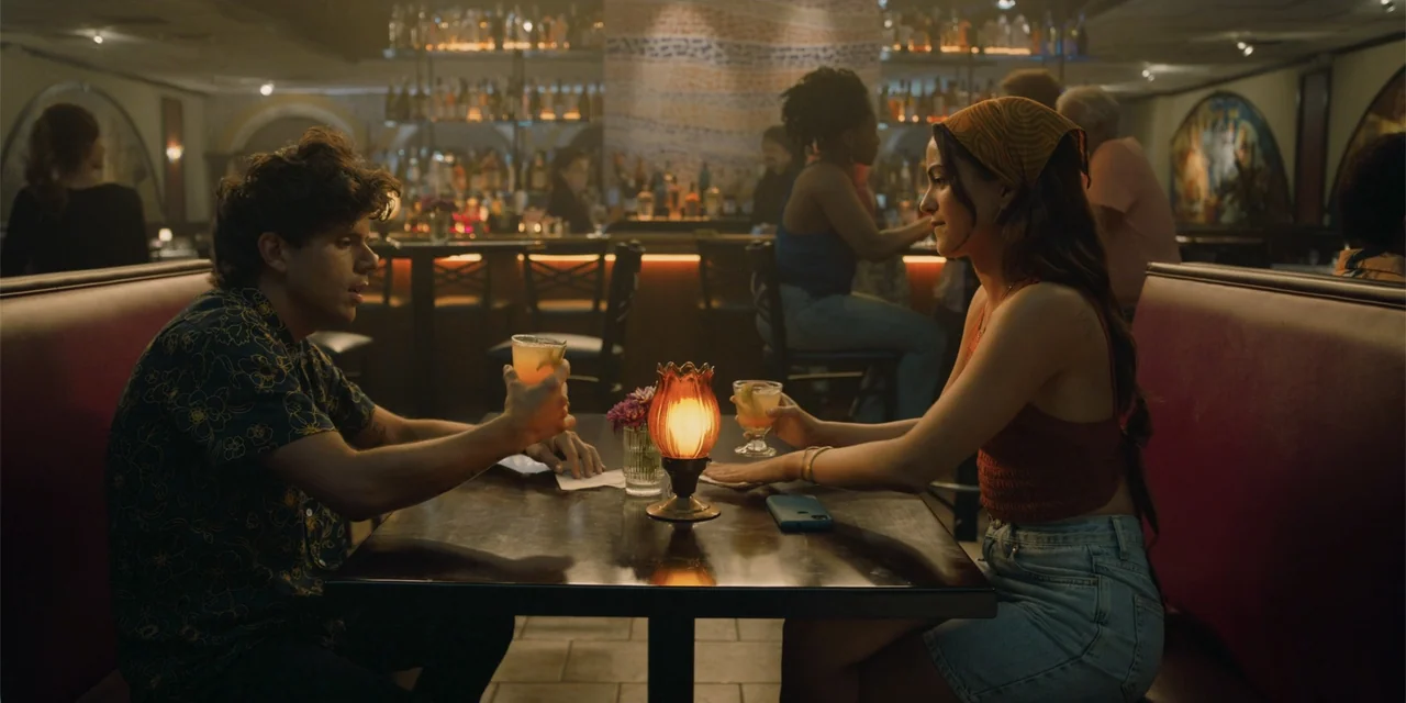 Música (2024) movie still: medium — A dark restaurant interior features red vinyl booths and a bar area in the background. A young man and woman sit…