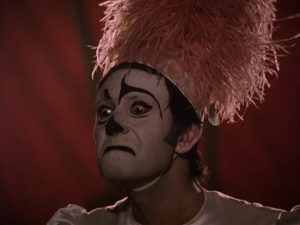 The Clowns (1970) movie still: closeup — A close-up of a mime wearing a pink feathered headpiece and dramatic white makeup. The performer stares…