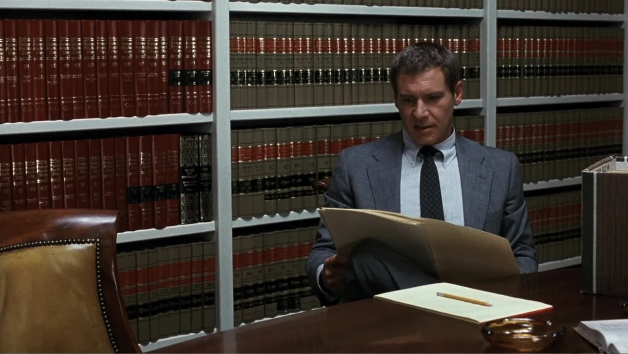 Presumed Innocent (1990) movie still: medium — A man sits at a wooden desk in front of shelves filled with books. He is reading a paper document…