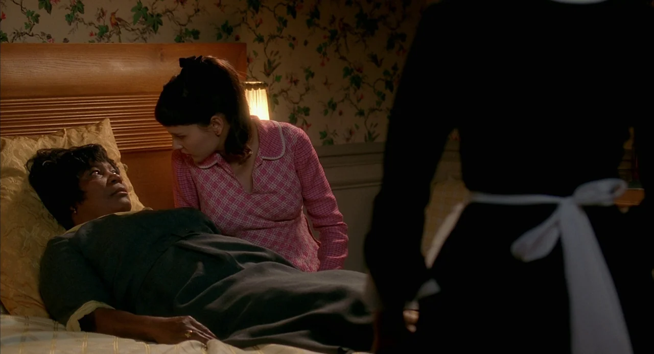 8 Women (2002) movie still: medium — A vintage bedroom features floral wallpaper and a wooden headboard with warm lamp illumination. A woman leans…