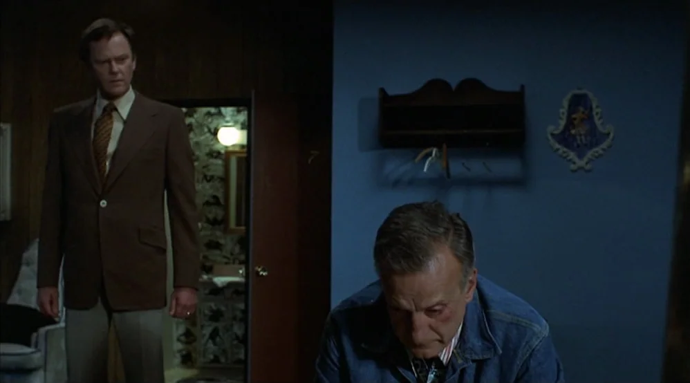 Hardcore (1979) movie still: medium — A blue-walled room features wood paneling and a doorway leading to a tiled bathroom. One man stands by the…