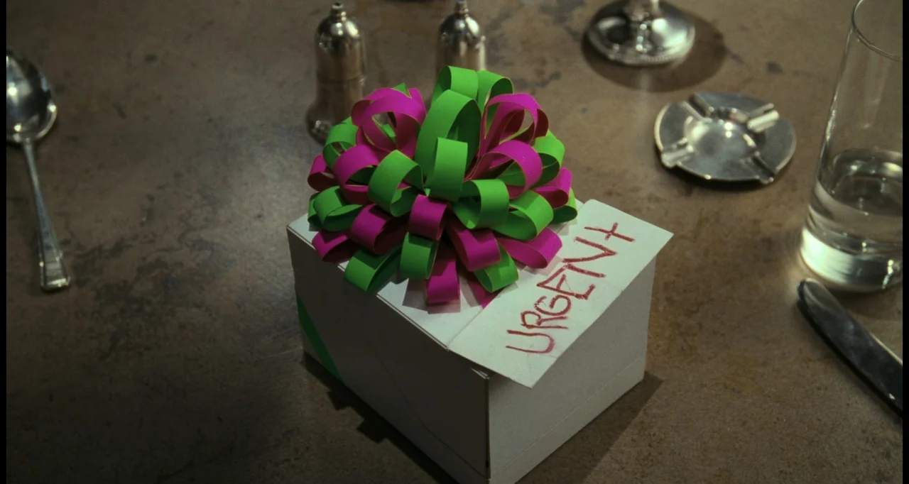 Batman (1989) movie still: closeup — A square gift box with a multi-colored bow rests on a textured brown surface. A hand-written card labeled…