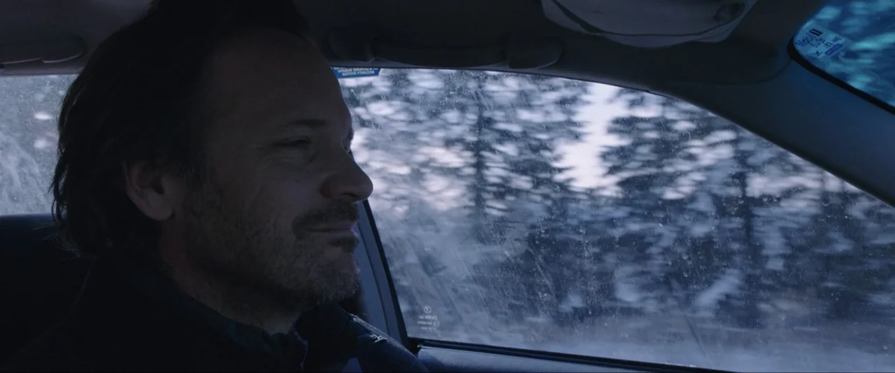 The Lie (2018) movie still: closeup — Inside a car cabin with a dirty side window facing a wintery forest. A man sits in the driver seat looking…