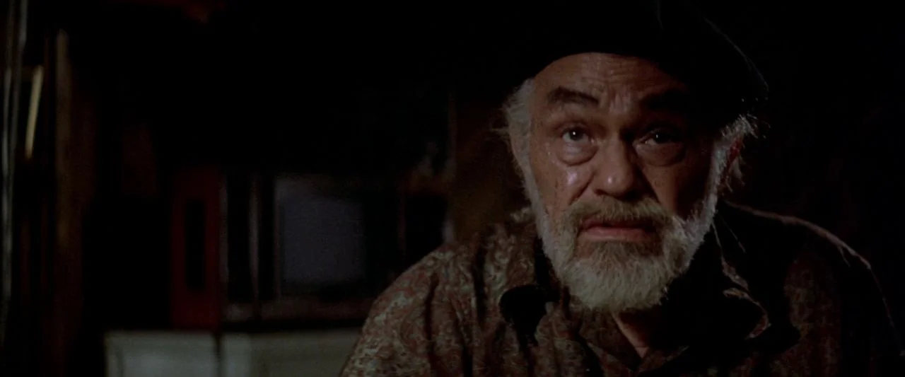 Soylent Green (1973) movie still: closeup — A dimly lit room with heavy shadows surrounding an elderly man in a beret. The man looks forward toward…