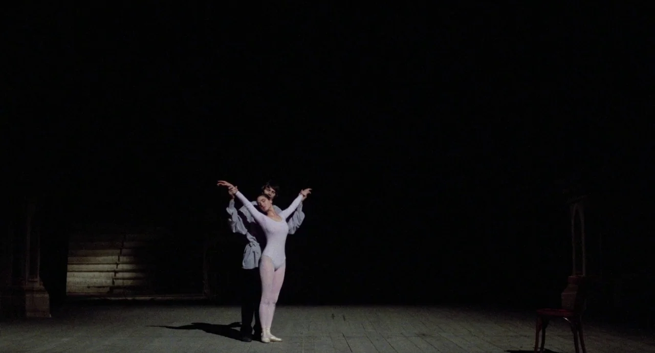 Etoile (1989) movie still: wide — A wide view of a dimly lit, empty wooden stage. A man and a woman in dance gear stand in a ballet pose under a…