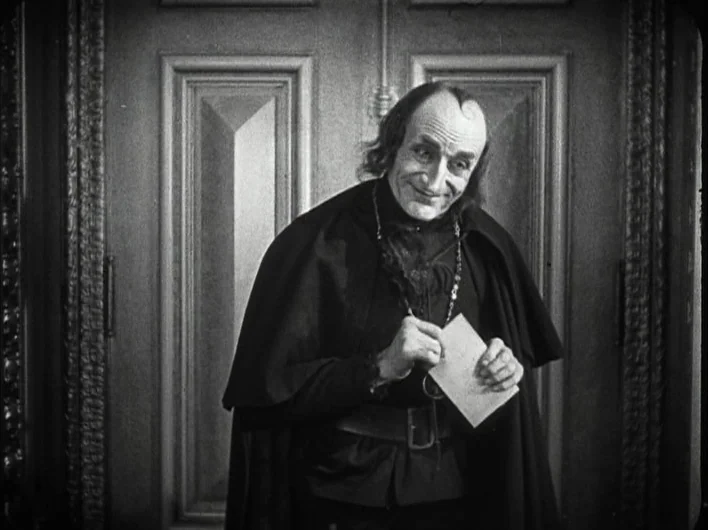 The Man Who Laughs (1928) movie still: medium — An elderly man in a dark cloak stands before a paneled wooden door. He clutches a white piece of…