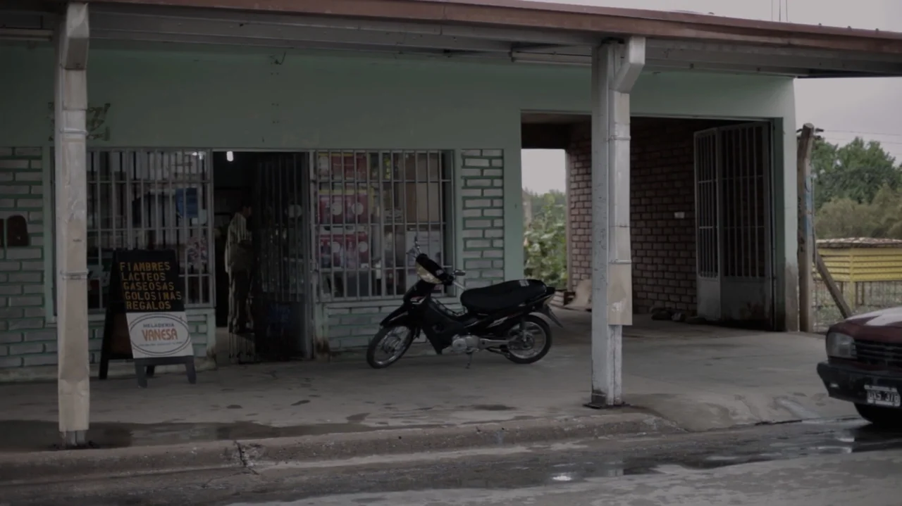 Still Life (2013) movie still: medium — A small concrete convenience store building with green walls and metal security grilles. A black motorcycle…