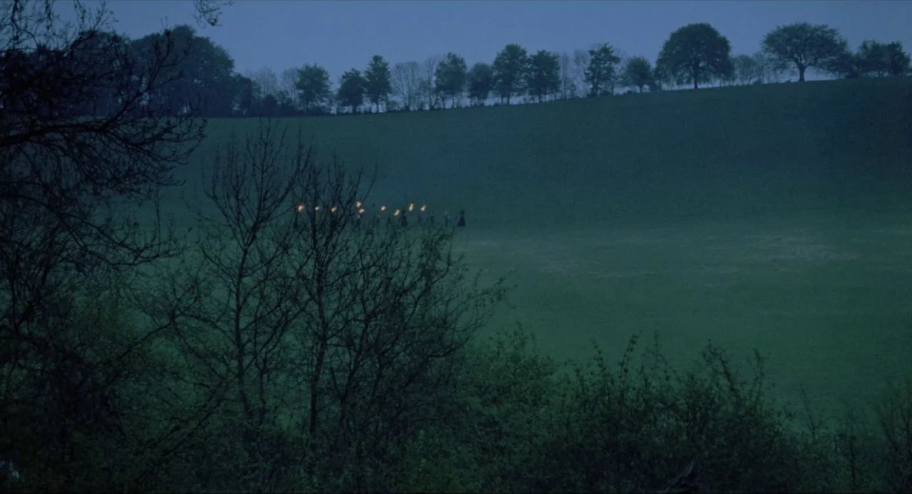 The Blood on Satan’s Claw (1971) movie still: wide — A vast, rolling green meadow is framed by dark trees during the blue hour. A small group of…
