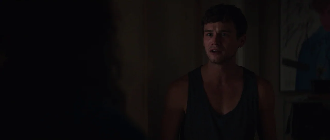 Hellraiser (2022) movie still: medium — A dimly lit room with shadows obscuring parts of the frame. A man in a tank top looks toward someone out of…