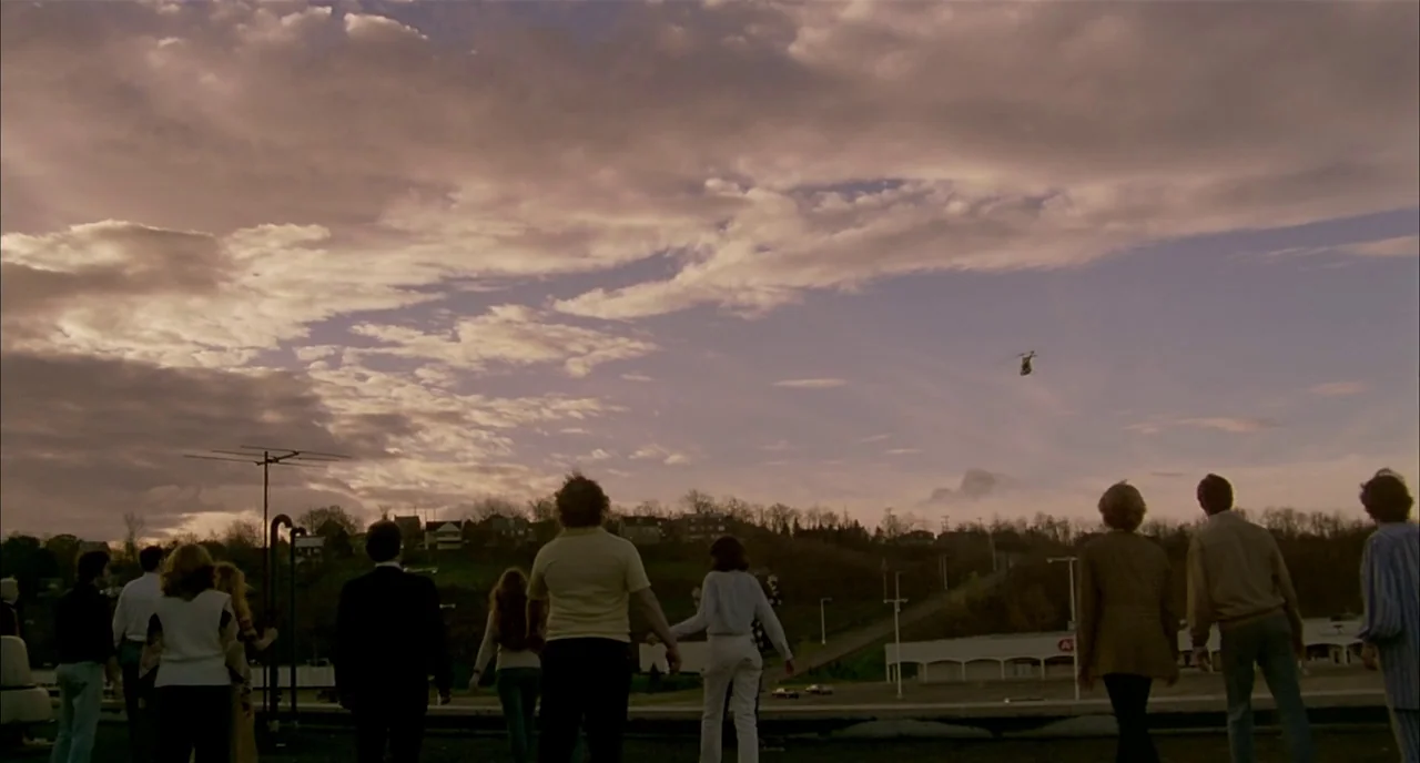Dawn of the Dead (1978) movie still: wide — A group of people stands on a flat rooftop facing a distant horizon at sunset. A small helicopter is…