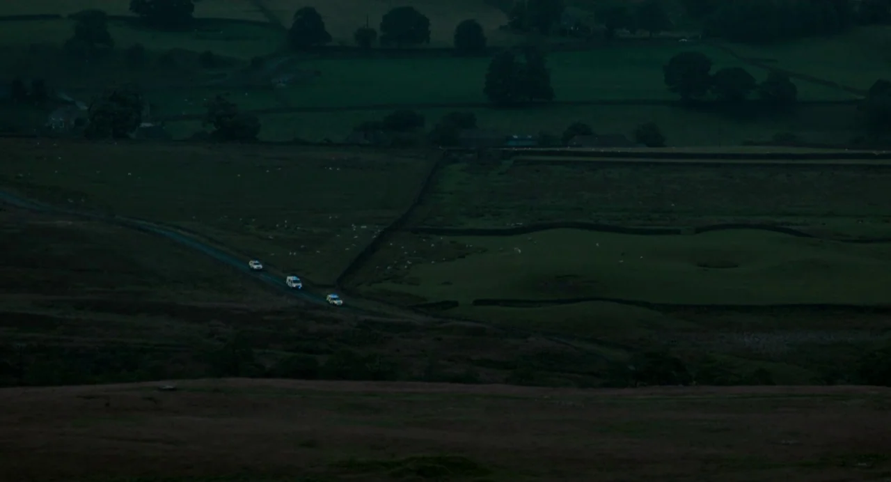 Dark River (2017) movie still: establishing — A vast, dark rural expanse of fields and stone walls sits under low light. Three small vehicles with…