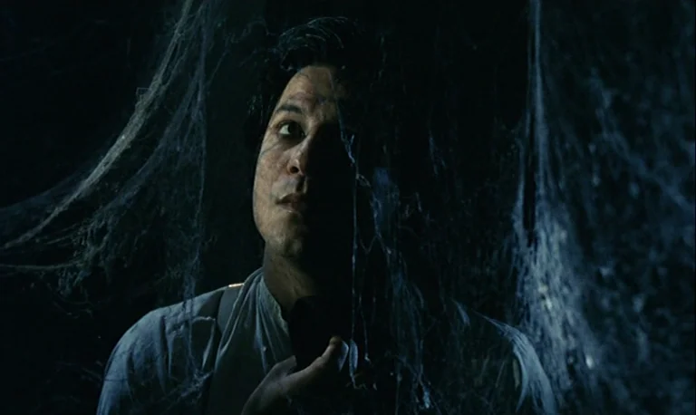 The Rose King (1986) movie still: closeup — A dark space covered in dense spiderwebs illuminated by a dim, low light. A man in a white shirt looks…