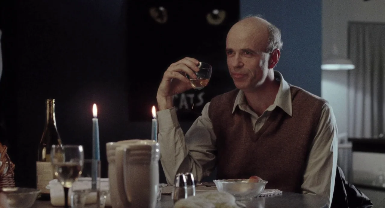 What Happened Was… (1994) movie still: medium — A dark dining room features a table set with a wine bottle and candles. A bald man in a sweater vest…