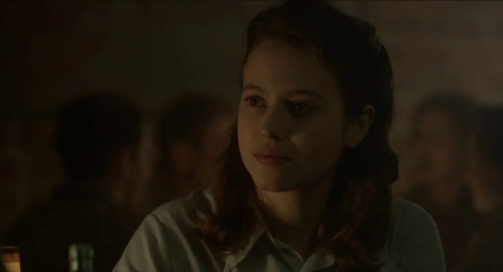 The Innocents (2016) movie still: closeup — A dimly lit, shadowed public house interior with out-of-focus patrons in the background. A young woman…