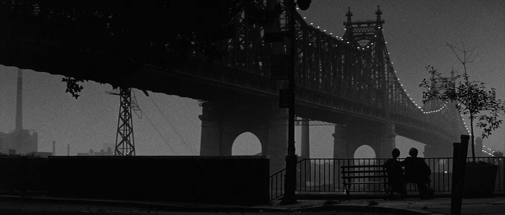 Manhattan (1979) movie still: wide — A night view across a railing toward a large industrial suspension bridge. Two figures sit on a public bench in…
