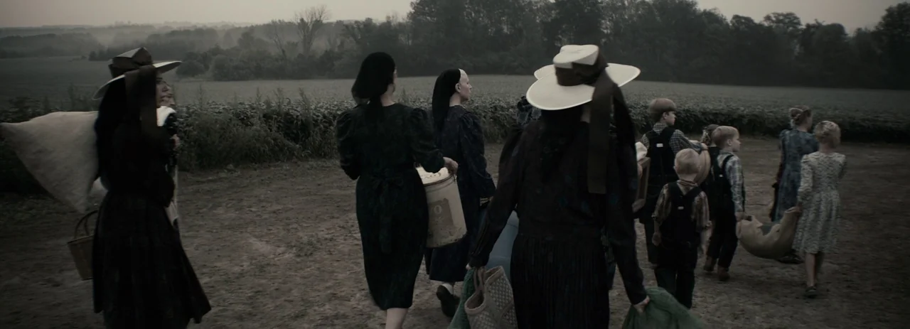 Women Talking (2022) movie still: wide — A group of people in 19th-century attire walks through a dusty rural landscape. They carry bags and buckets…