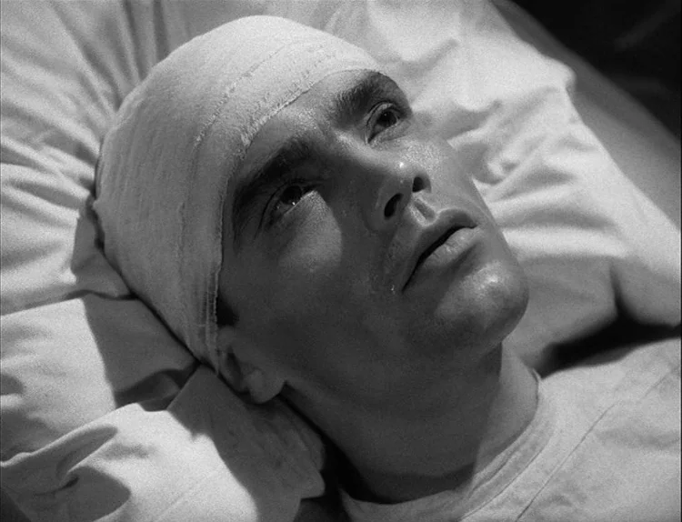 Music in Darkness (1948) movie still: closeup — A man lies on white pillows wearing a bandage around his head. He gazes upward from his position in…