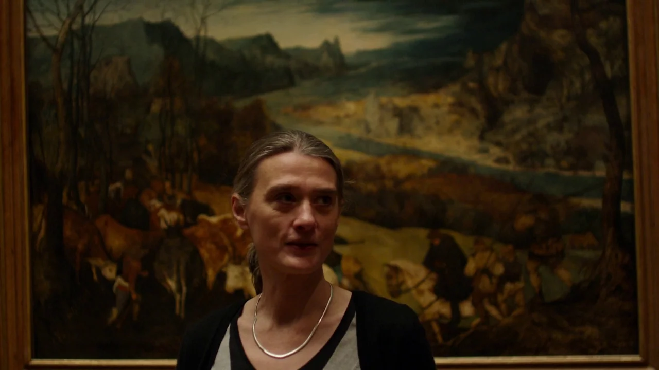 Museum Hours (2012) movie still: medium — A woman stands in a museum gallery in front of a framed landscape painting. The painting depicts a…
