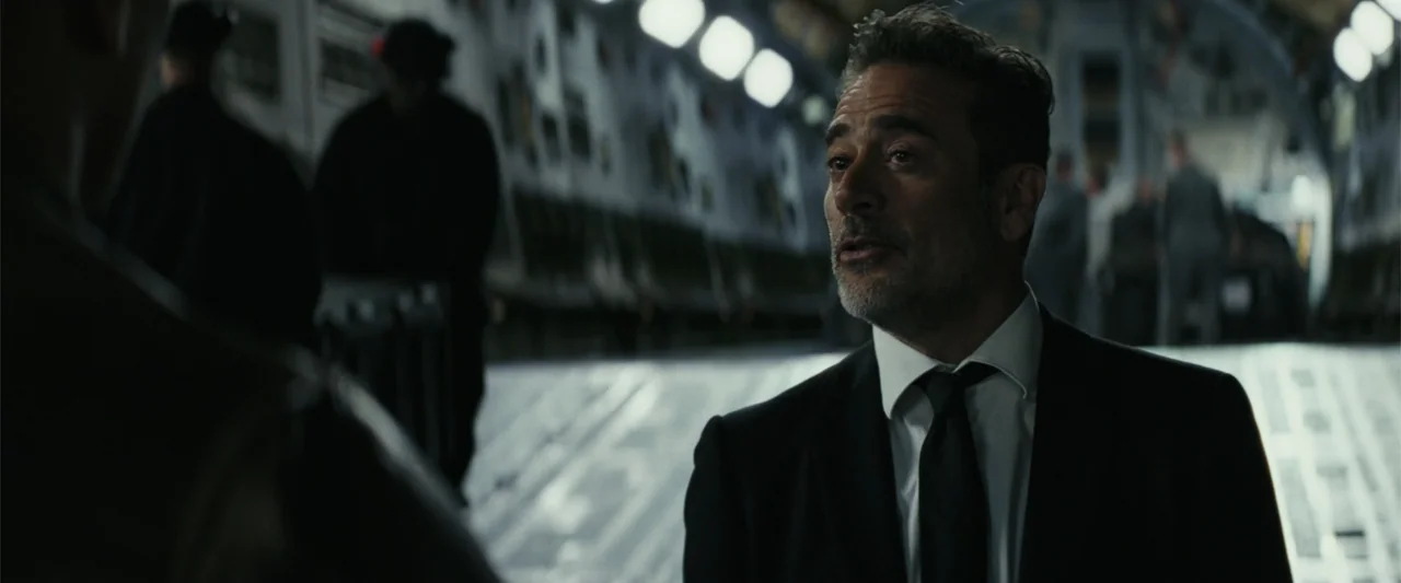 Rampage (2018) movie still: medium — A man stands inside the cargo hold of an airplane with blurred figures behind. The man wearing a suit looks…