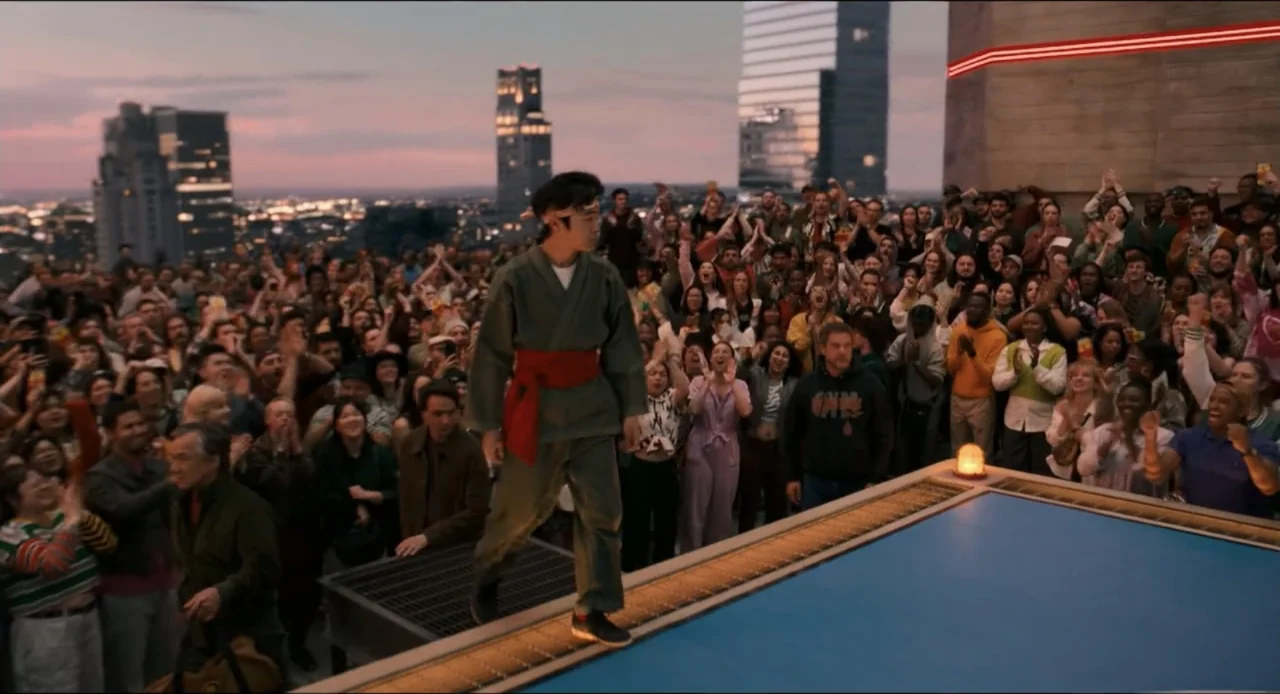 Karate Kid: Legends (2025) movie still: fullbody — A rooftop platform overlooking a city skyline at sunset with a large crowd cheering. A young man…