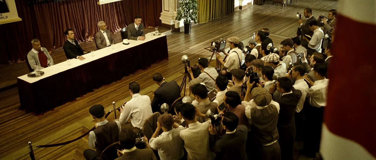 Ip Man 2 (2010) movie still: wide — A spacious wood-floored hall with long red-draped tables arranged for a formal presentation. A crowd of…