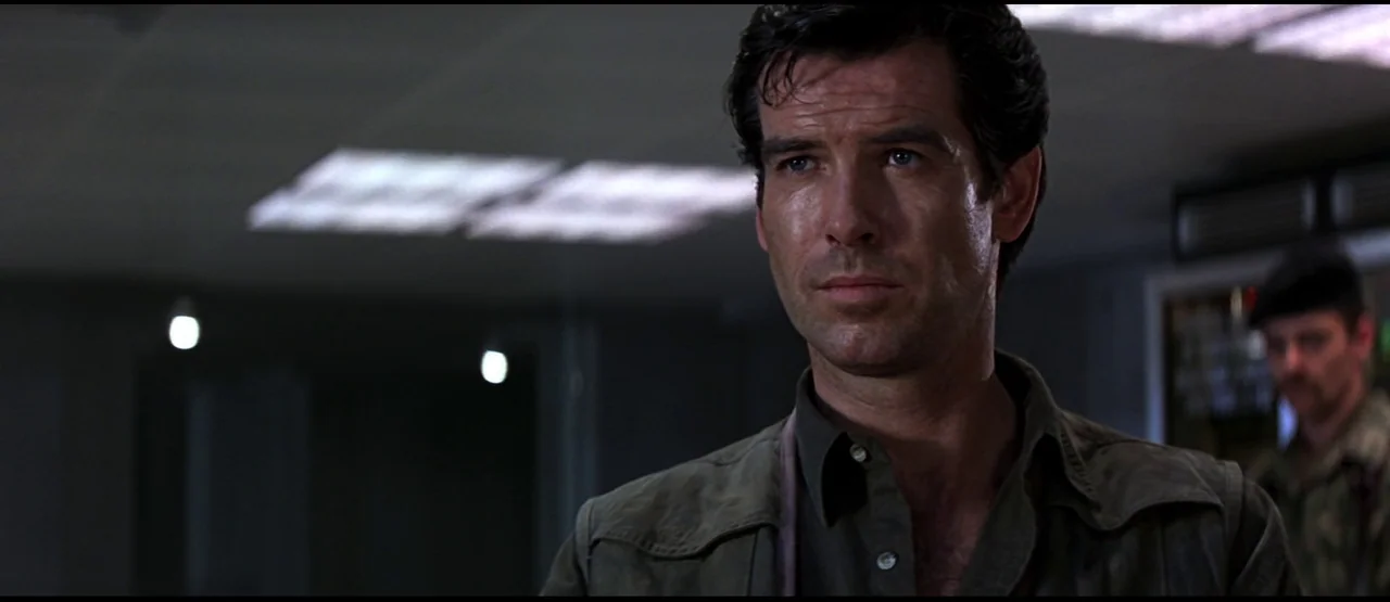 GoldenEye (1995) movie still: closeup — A dimly lit room features a person with a short dark haircut in front. The background includes a second…