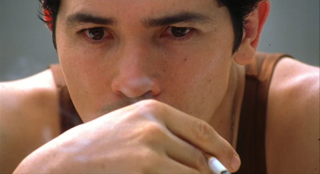 Summer of Sam (1999) movie still: closeup — A man in a brown tank top is indoors during the day. He holds a cigarette near his mouth while looking…