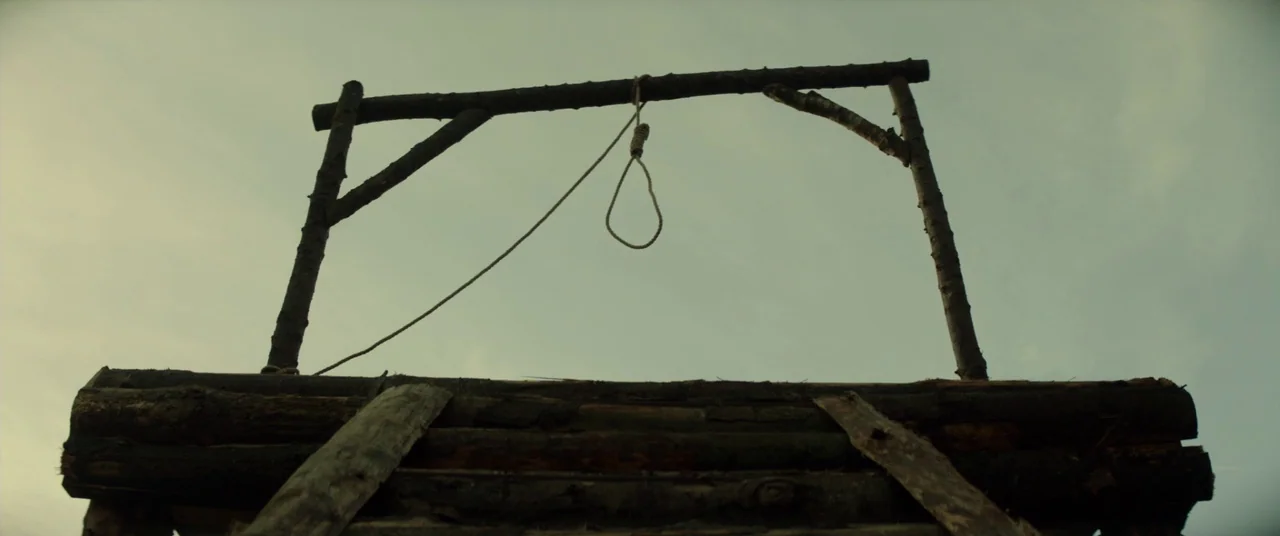 Never Grow Old (2019) movie still: fullbody — A wooden gallows structure stands against an overcast sky in an open area. The frame shows the…