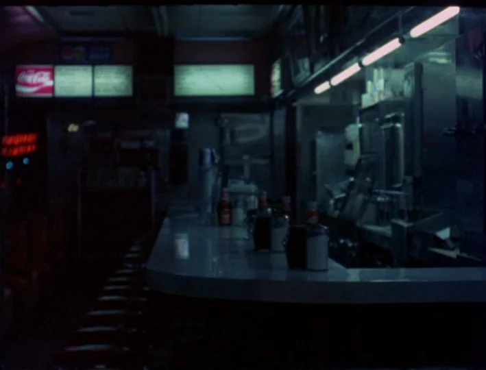News From Home (1977) movie still: wide — A dimly lit 1970s style diner with an empty curved counter and polished surface. Rows of dark stools line…