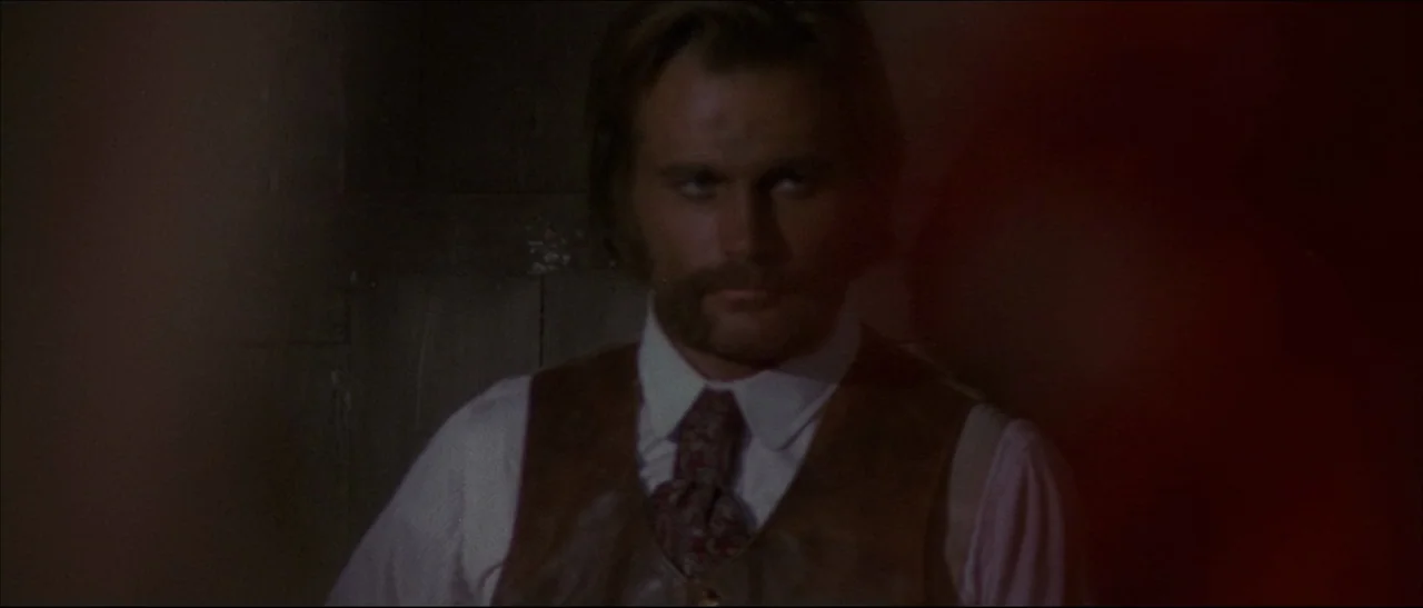 The Mercenary (1968) movie still: medium — A dimly lit interior space features rough-hewn wooden walls and blurred foreground obstructions. The man…