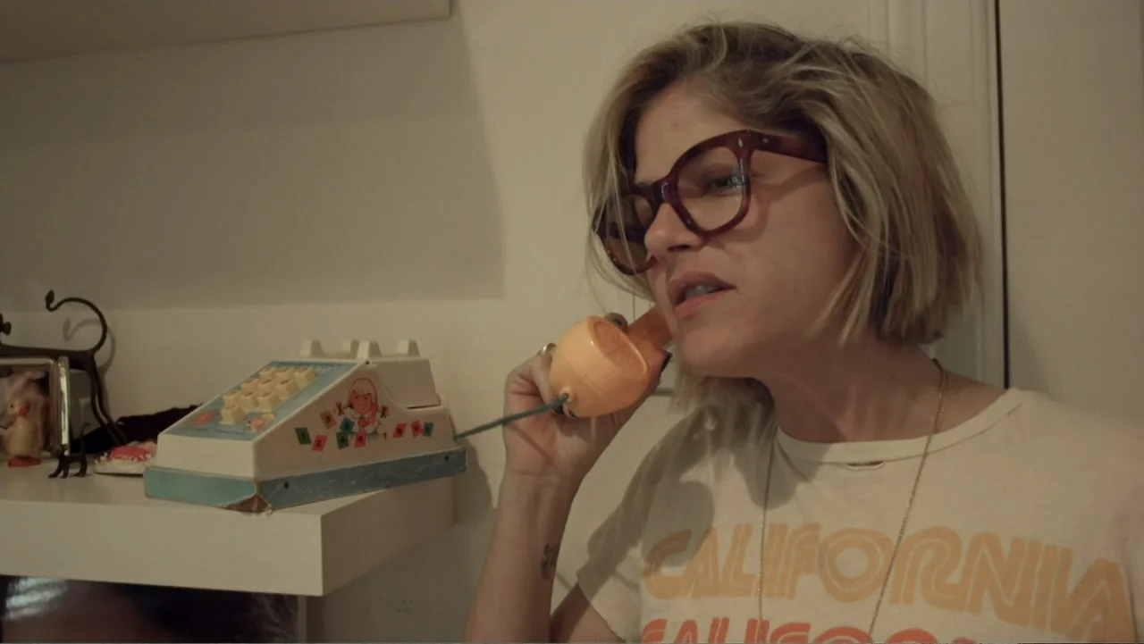 Introducing, Selma Blair (2021) movie still: medium — An interior wall features a white shelf holding a retro toy telephone. A blonde woman with…
