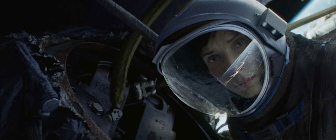 Gravity (2013) movie still: closeup — A cramped spacecraft cockpit with exposed wiring and metallic panels surrounding a human occupant. An…
