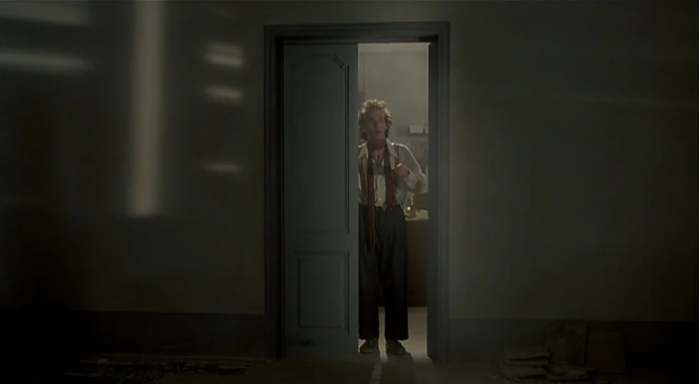The Voice of the Moon (1990) movie still: fullbody — A dimly lit room faces an open door leading to a brighter interior space. A man in a light…