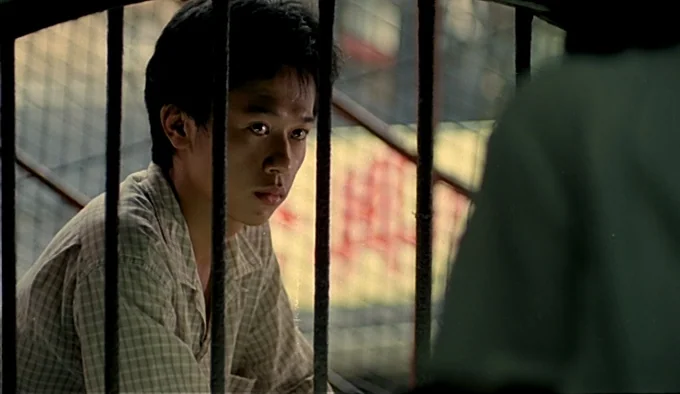 Dust In The Wind (1986) movie still: medium — A man sits behind thick, dark vertical metal bars in an indoor space. The man looks toward an unseen…