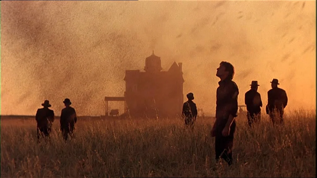 Days Of Heaven (1978) movie still: wide — A field of tall grass sits under an orange sky during golden hour. Several men stand scattered in the…