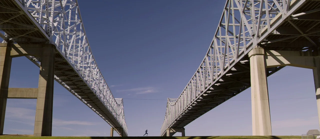 Self/less (2015) movie still: wide — A massive steel truss bridge structure towers over a grassy horizon under clear sky. A small figure in…
