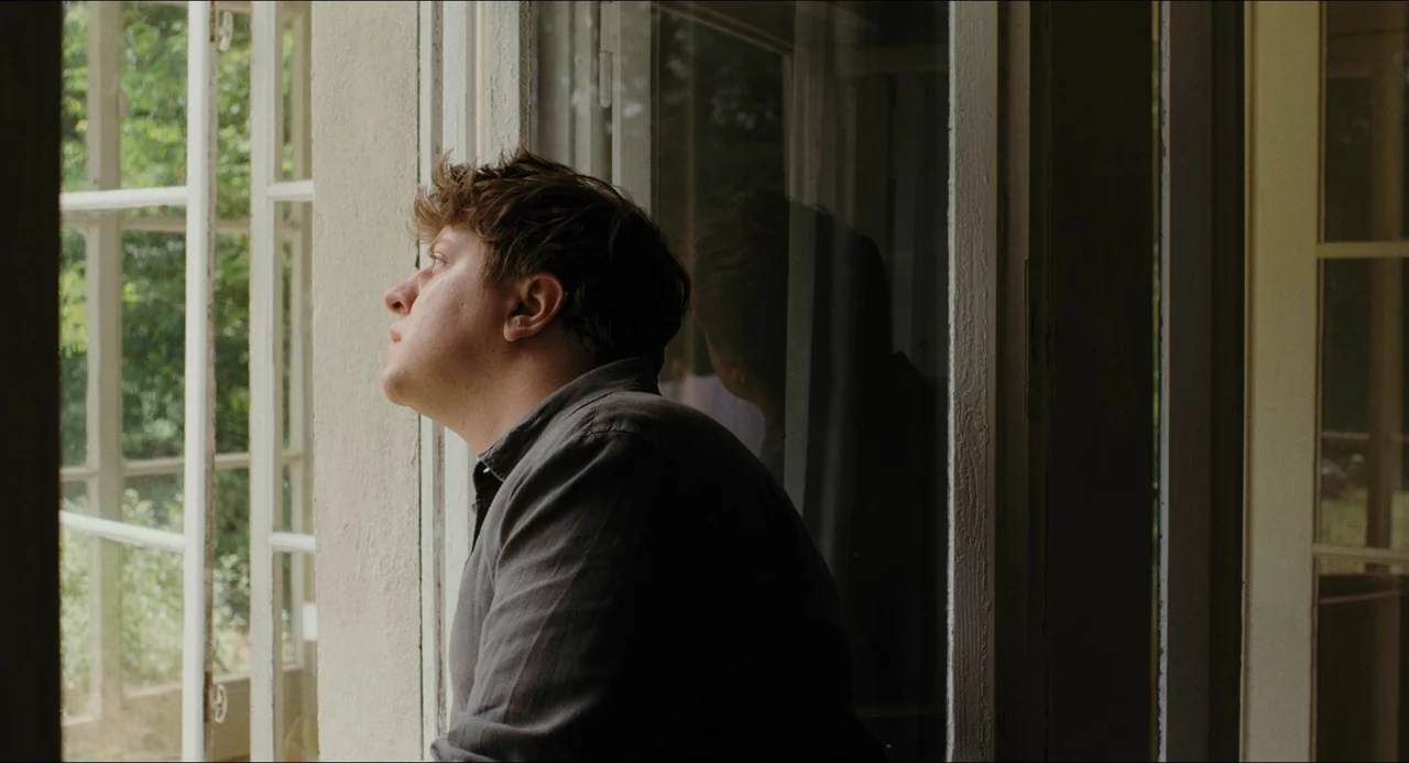 Afire (2023) movie still: medium — An interior space with a large open window looking out onto green foliage. A man stands at the window gazing…