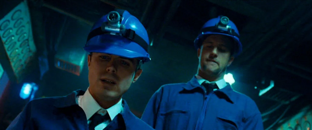 Ocean’s Thirteen (2007) movie still: medium — Two men stand inside a dim, industrial environment with visible overhead machinery and pipes. They…
