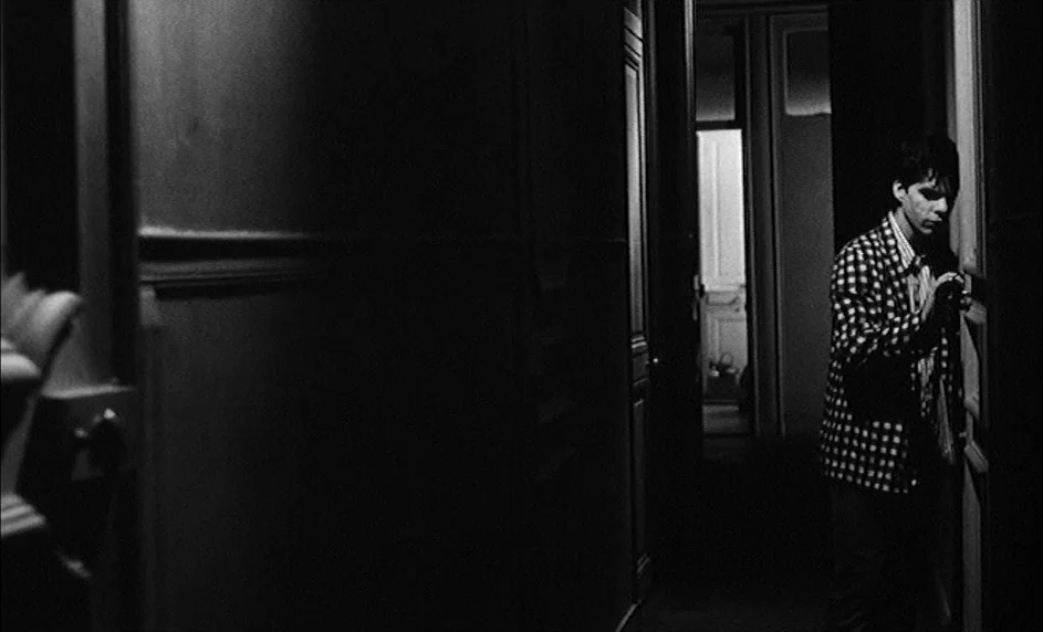 Boy Meets Girl (1984) movie still: medium — A dark, narrow domestic hallway with paneled walls features a single door in view. A young man in a…