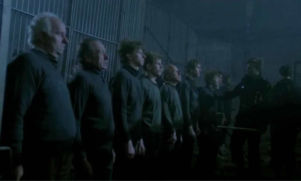 Ga-ga: Glory to the Heroes (1986) movie still: fullbody — A dark industrial hall with metal fencing and concrete walls defines the space. A line of…